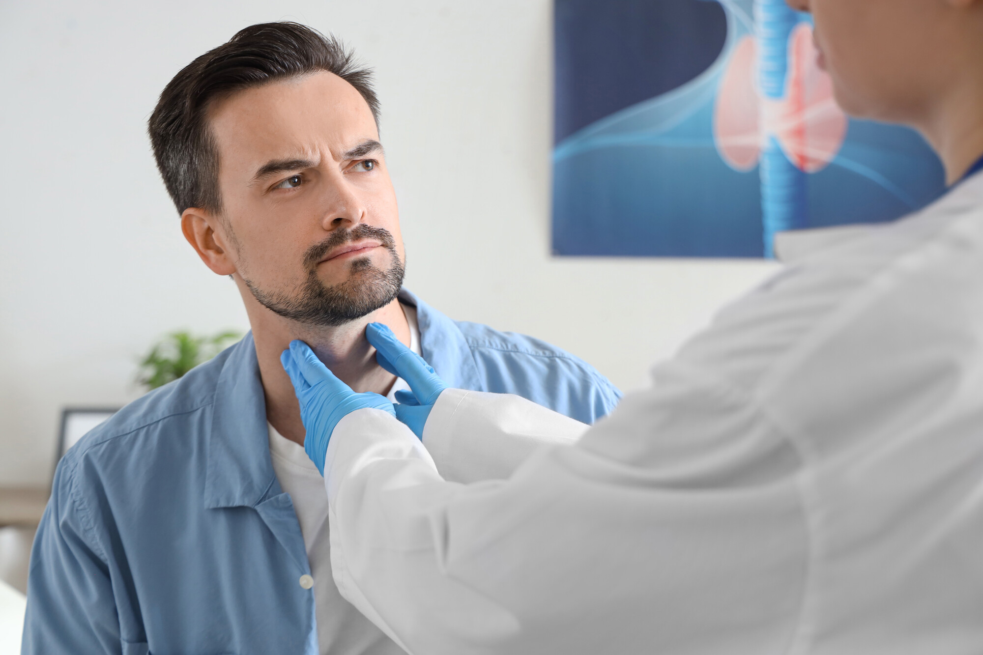 Medico in camice e guanti blu palpa il collo di un uomo barbuto in camicia azzurra, poster della tiroide sullo sfondo